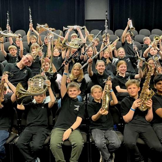 En stor gruppe sidder med deres blæseinstrumenter i en koncertsal