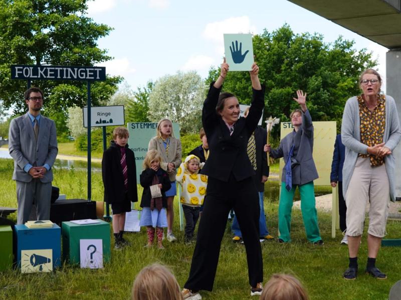 En gruppe skuespillere står udenfor med byskilte og stemmebokse som rekvisitter. 
